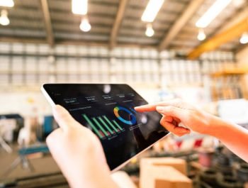 Action of a worker is using tablet to review storage report with blurred background of warehouse. Business management and technology concept photo. Close-up and selective focus at human's hand part.